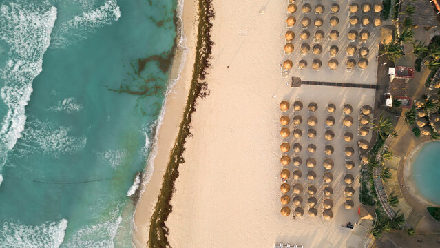 Aerial view of the beach with rows of straw umbrellas on white sand, turquoise Caribbean sea waves, and a swimming pool area in Playa del Carmen, Quintana Roo, Mexico.