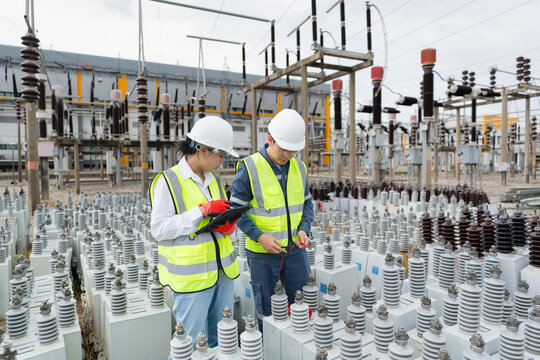 In a substation switchyard, capacitor unit inspection involves two technicians examining an insulator with a tablet beside a steel gantry.