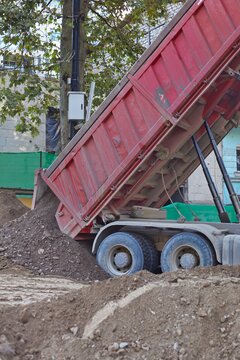 Red dump truck unloading soil and gravel at city road construction site
