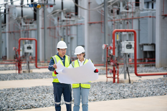 Power transformer inspection at the substation proceeds with two engineers in hard hats reviewing a blueprint by the control cabinet.
