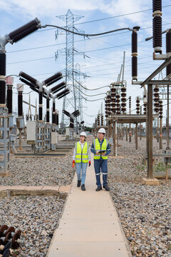 Two technicians patrol a substation inspection route, walking with a tablet beside a high voltage disconnect switch and lattice tower.