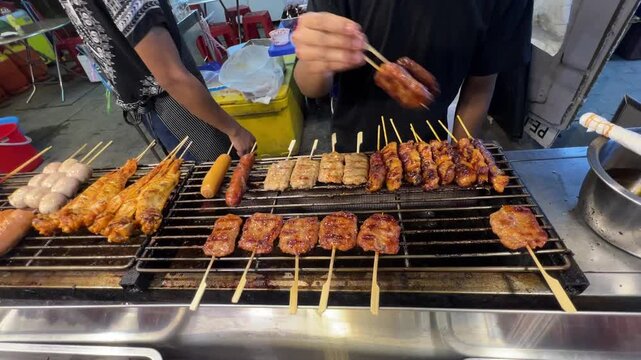 Street food vendor grilling assorted meat skewers, chicken, sausages and meatballs over barbecue grill at a busy Asian market, showcasing freshly cooked snacks, local cuisine and outdoor food culture.