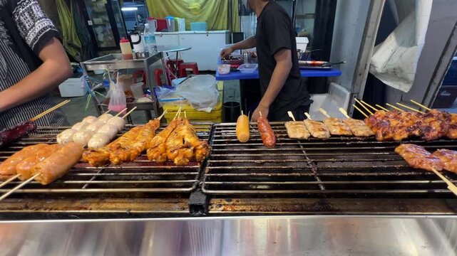 Street food vendor grilling assorted meat skewers, chicken, sausages and meatballs over barbecue grill at a busy Asian market, showcasing freshly cooked snacks, local cuisine and outdoor food culture.