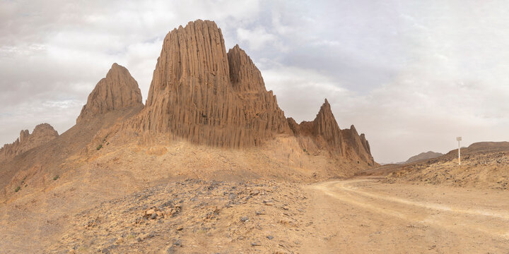 Towering volcanic rock spires with vertical organ-pipe fluting stand along an unpaved desert dirt road in the Hoggar Mountains near Assekrem, Tamanrasset, Algeria under an overcast sky.
