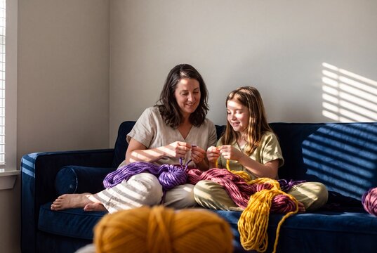 Smiling mother and daughter sitting on sofa knitting together with colorful chunky yarn at home in sunlight