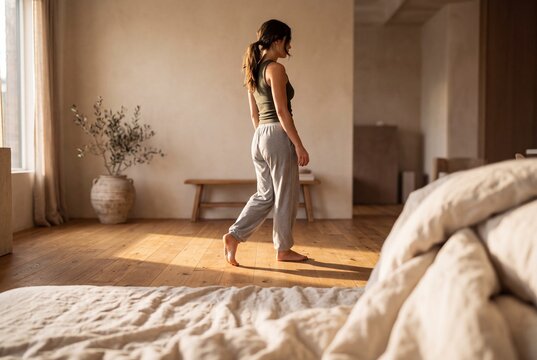 Young woman in comfortable loungewear walking barefoot in a bright minimalist bedroom filled with warm morning sunlight