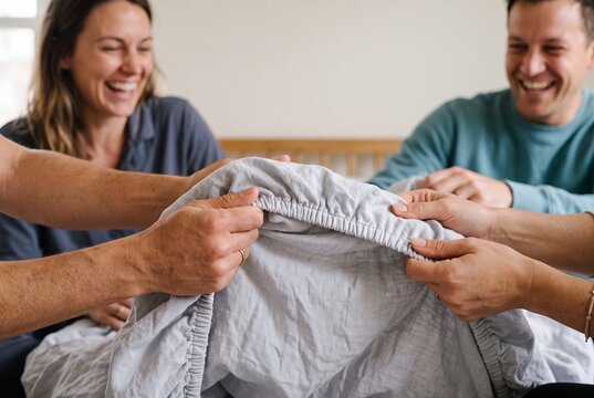 Happy family and couple laughing together while struggling to fold a fitted bed sheet in a bright bedroom