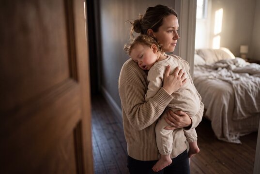 A tired young mother holding her sleeping baby in her arms while standing near the bedroom doorway at home