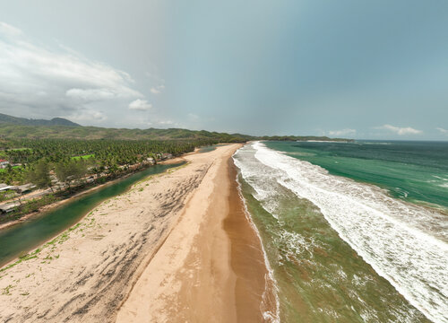 Aerial view of Soge Beach with a river flowing parallel to the sandy coastline, lush palm trees, and rolling hills in Kecamatan Ngadirojo, Jawa Timur, Indonesia.