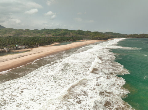 Aerial view of Soge Beach with white sand, turquoise waves, and lush green hills under a cloudy sky in Kecamatan Ngadirojo, Jawa Timur, Indonesia.