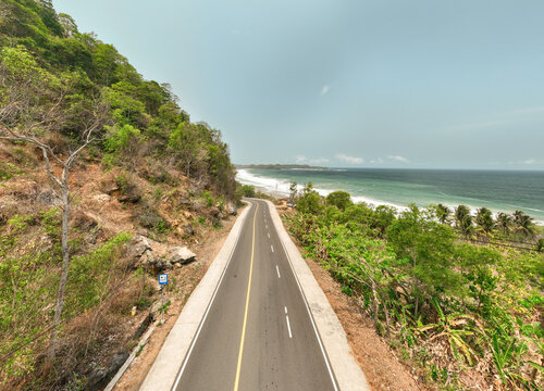 Aerial view of Soge Beach with a coastal road winding along the cliffside, lush green vegetation, and ocean waves in Kecamatan Ngadirojo, Jawa Timur, Indonesia.