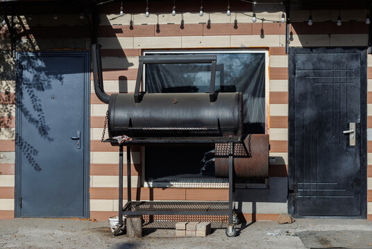 Professional heavy duty metal offset smoker grill standing outdoors near a striped wall with doors and string lights on a sunny day