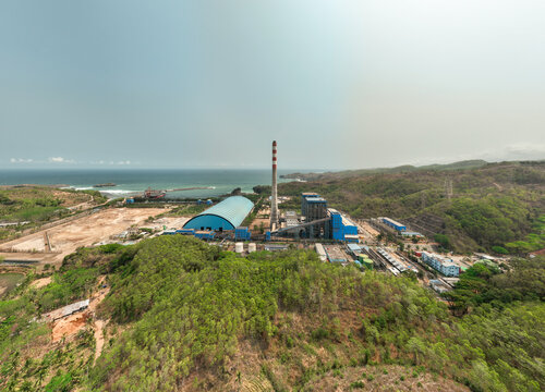 Aerial view of Pacitan Power Plant with its tall chimney and blue industrial buildings situated along the coastline near green hills in Kecamatan Sudimoro, Jawa Timur, Indonesia.