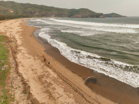 Aerial view of Pacitan Horseman riding horses along the sandy beach with rolling waves and lush hills in the background in Pacitan, East Java, Indonesia.