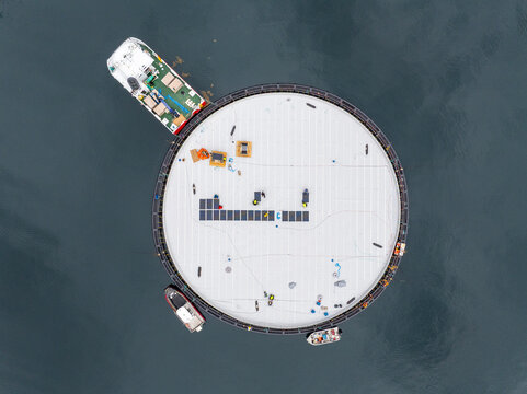 Aerial view of installation of solar panels on a marine fish farming ring with support vessels in the dark water of a fjord, Fjord, More og Romsdal, Norway.