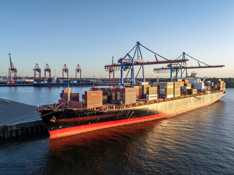 Aerial view of a large container ship docked at the Port of Hamburg with colorful shipping containers and gantry cranes under clear skies in Hamburg, Hamburg, Germany.