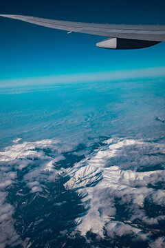 Aerial view of snow-capped mountains and clouds seen from an airplane window with the wing visible against a clear blue sky Oslo, Oslo, Norway.