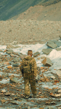 Hiker in camouflage gear walking across rocky mountain terrain with patches of snow and distant green slopes