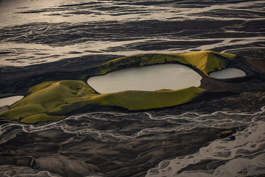 Aerial view of vibrant green moss-covered volcanic craters and glacial lakes of Landmannalaugar set against dark volcanic sand and braided river patterns in Hella, Rangarthing ytra, Iceland.
