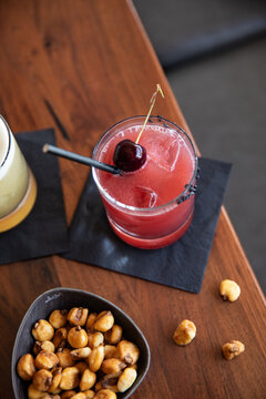craft cocktail garnished with cherry on black napkin on wood table with fried corn kernels as snack in upscale bar