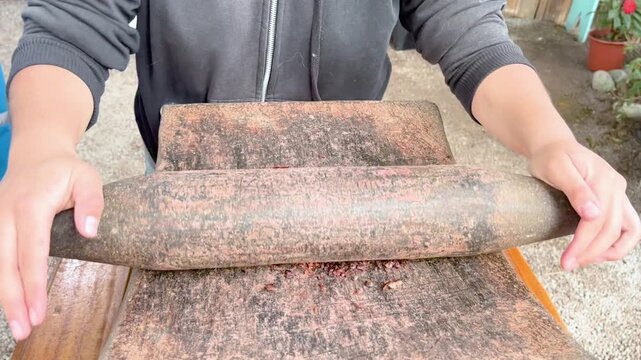 A person grinding roasted cocoa beans by hand using a traditional stone rolling pin on a stone surface, demonstrating the artisanal method of cocoa processing and food production techniques.