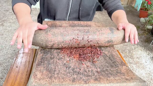 A person grinding roasted cocoa beans by hand using a traditional stone rolling pin on a stone surface, demonstrating the artisanal method of cocoa processing and food production techniques.