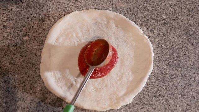 Overhead slow motion of hand ladling and spreading tomato sauce onto pizza dough on kitchen countertop, top down tracking shot.
