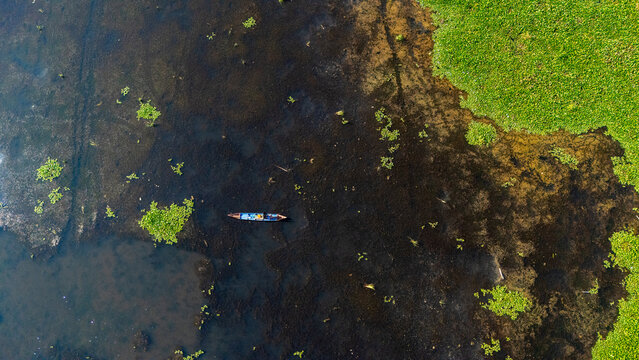 Aerial view of a small boat navigating through dark water with patches of green aquatic vegetation in Salatiga, Central Java, Indonesia.