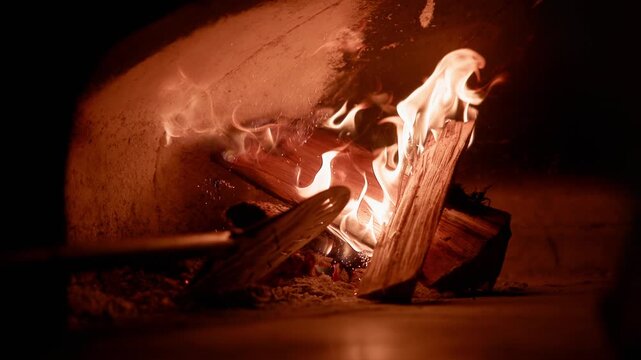 Tracking shot of peel stoking burning wood logs and dancing flames inside brick oven hearth, low closeup view showing embers and flickering orange glow in pizzeria.