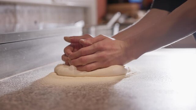 Tracking shot hands shaping pizza dough on floured countertop in commercial kitchen, close up movement.
