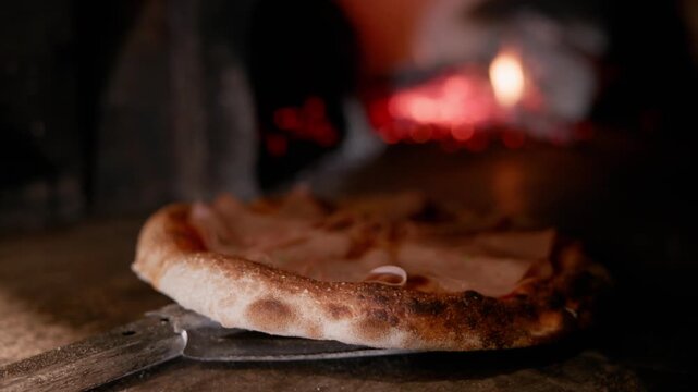 Tracking shot of pizza sliding out of wood fired oven on peel in rustic kitchen close up of charred crust and melting cheese.