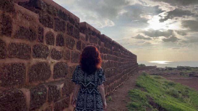 Tracking shot of woman with red hair walking along the laterite wall of Chapora Fort in Goa towards the sunset sea