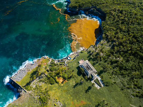 Aerial view of a coastal building perched on a cliff edge overlooking turquoise waters and a patch of sargassum seaweed near Las Galeras, Samana, Samana Province, Dominican Republic.
