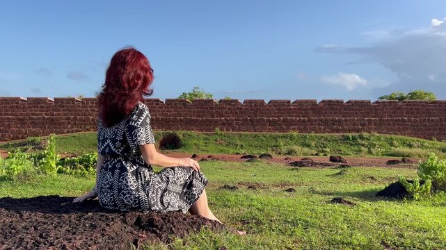 Static shot of woman with red hair sitting on stone, looking at the laterite wall of Chapora Fort in Goa