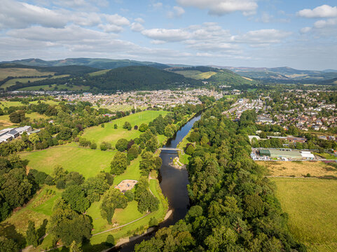 Aerial view of the town of Peebles along the River Tweed, featuring green fields, dense woodland, and rolling hills under a bright sky in Peebles, Scotland, United Kingdom.