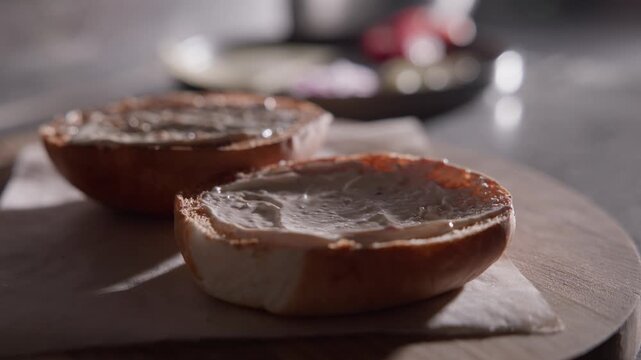 Slow motion close up of toasted bagel halves spread with cream cheese on wooden cutting board on kitchen countertop.