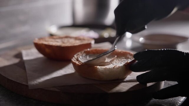 Slow motion close up of gloved hand spreading jam on burger brioche bun on wooden cutting board in kitchen.