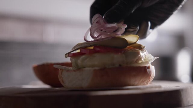 Closeup slow motion placing pickles and sliced onions onto cheeseburger on cutting board in kitchen.