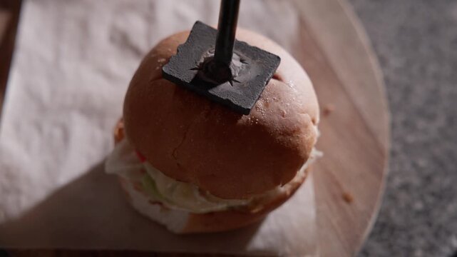 Closeup burger being pressed with metal tamper on cutting board and parchment on kitchen countertop.
