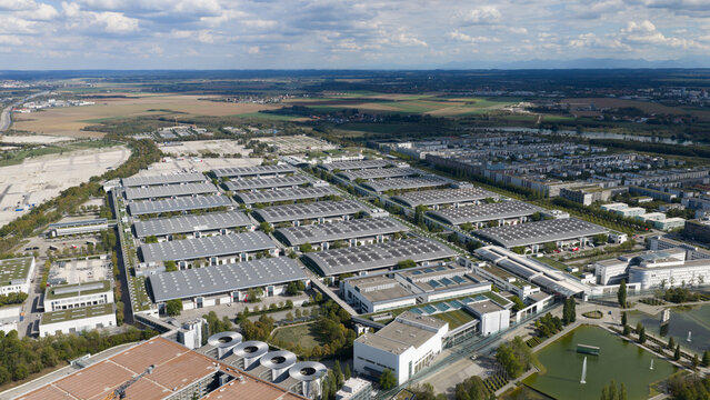 Aerial view of Messe Munchen exhibition center with large interconnected halls, flat roofs, surrounding green spaces, and a nearby pond in Munchen, Bayern, Germany.