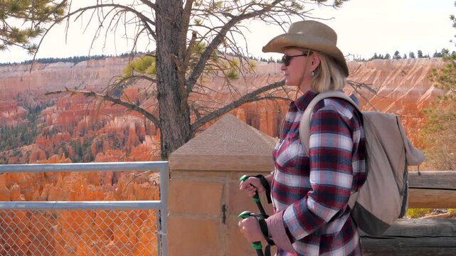 Mature woman with backpack and hiking poles walking along viewpoint. Hiker admiring the stunning orange hoodoos and geological formations of bryce canyon national park in utah