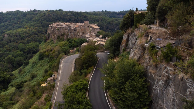 Aerial view of the historic village of Calcata Vecchia perched on a volcanic cliff surrounded by lush green forests and a winding road, Calcata Vecchia, Lazio, Italy.