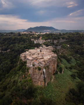 Aerial view of the historic village of Calcata Vecchia perched on a volcanic cliff surrounded by lush forests and rolling hills under a dramatic sky in Calcata Vecchia, Lazio, Italy.