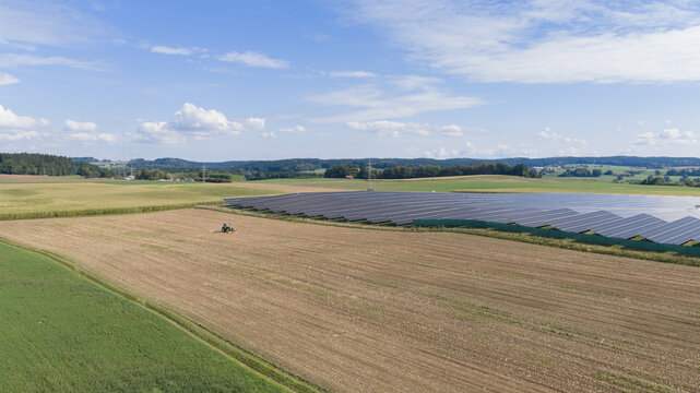 Aerial view of VISPIRON Solarpark Chargingstation with rows of photovoltaic panels in a rural landscape with agricultural fields and a tractor working nearby. Egling, Bavaria, Germany.