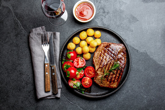 Grilled beef steak served on cast iron plate with tomato salad, potatoes balls and red wine. Barbecue, bbq meat beef tenderloin. Top view, slate background