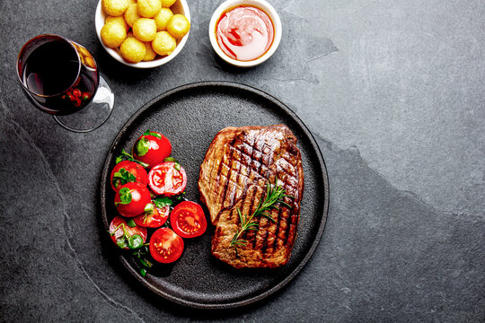 Grilled beef steak served on cast iron plate with tomato salad, potatoes balls and red wine. Barbecue, bbq meat beef tenderloin. Top view, slate background