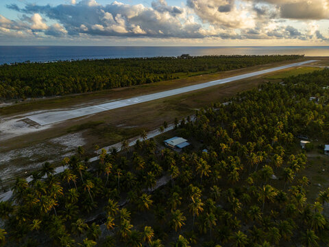 Aerial view of Tikehau airport runway surrounded by lush palm trees and tropical vegetation under a golden hour sky in The Tuamotu and Gambier Islands, French Polynesia.