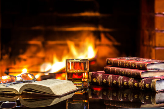Glass alcoholic drink with antique books in front warm fireplace