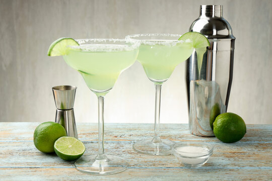 Refreshing margarita cocktail in glasses, limes, shaker and jigger on wooden table, closeup