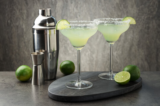 Refreshing margarita cocktail in glasses, limes, shaker and jigger on grey table, closeup